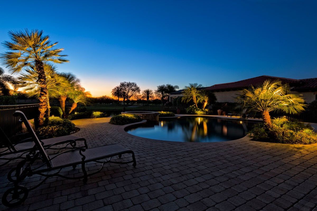 Nighttime view of a pool with patio, palm trees illuminated by lights, and a darkening sky.