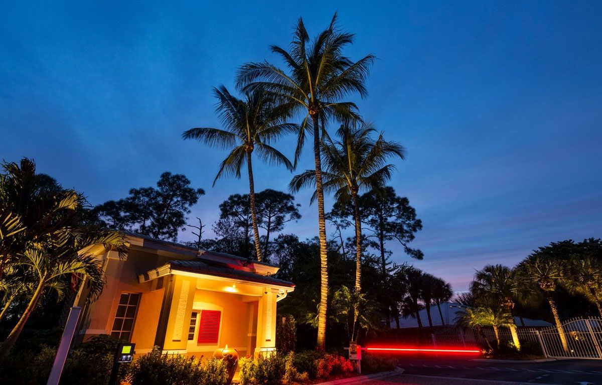 Entrance gate at dusk with lit building, palm trees, and blurred car lights.