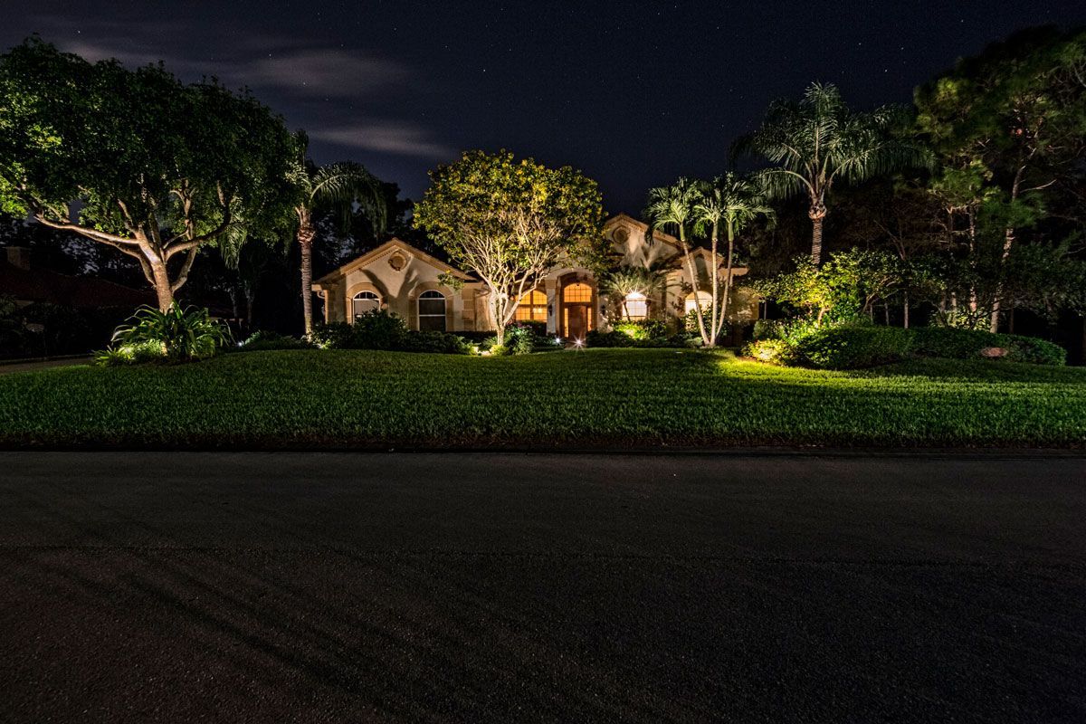 Night view of a house with landscape lighting illuminating the facade, trees, and lawn along a dark road.