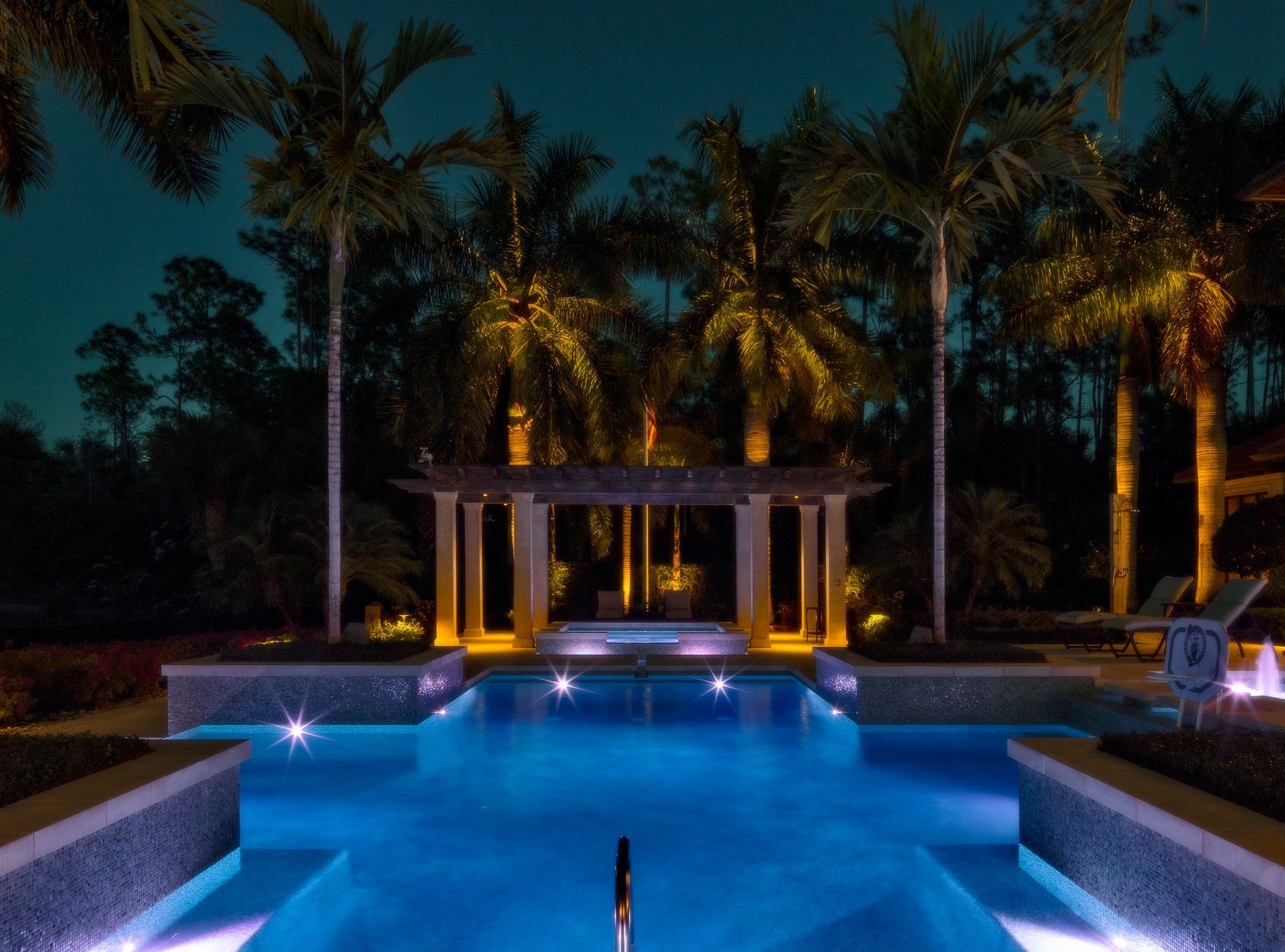 Nighttime pool with illuminated palm trees, pergola, and blue water.