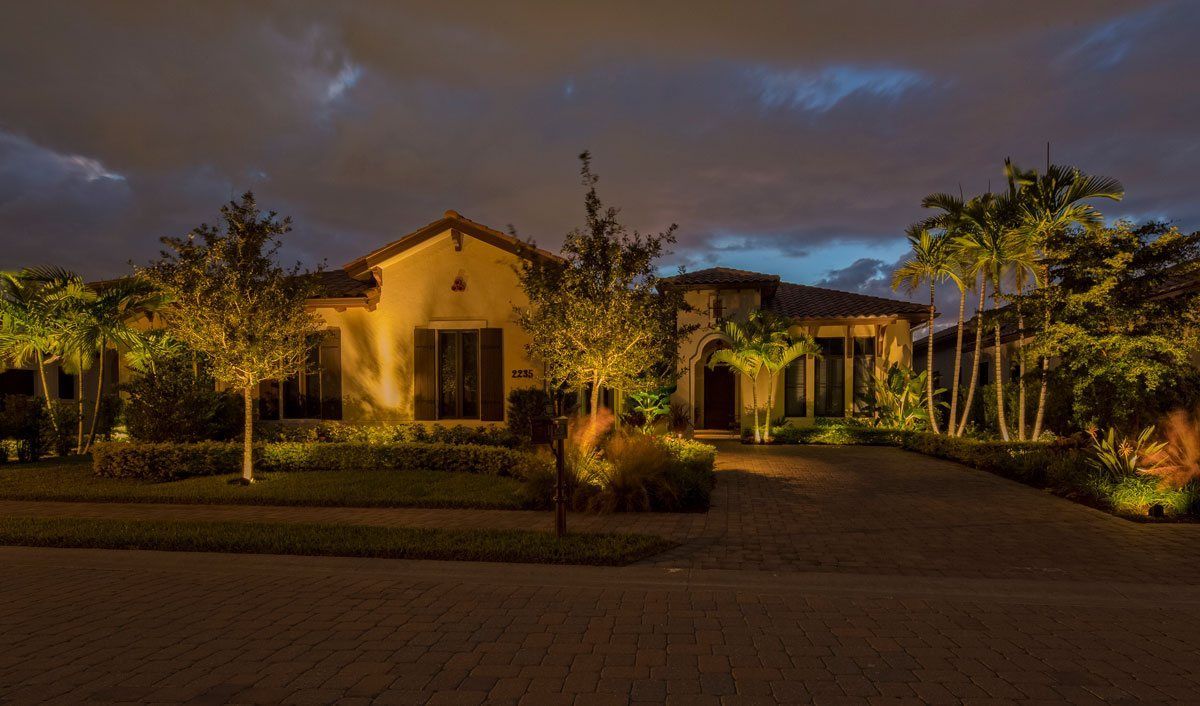 A lit house at dusk with trees, palm trees, and a driveway against a cloudy sky.