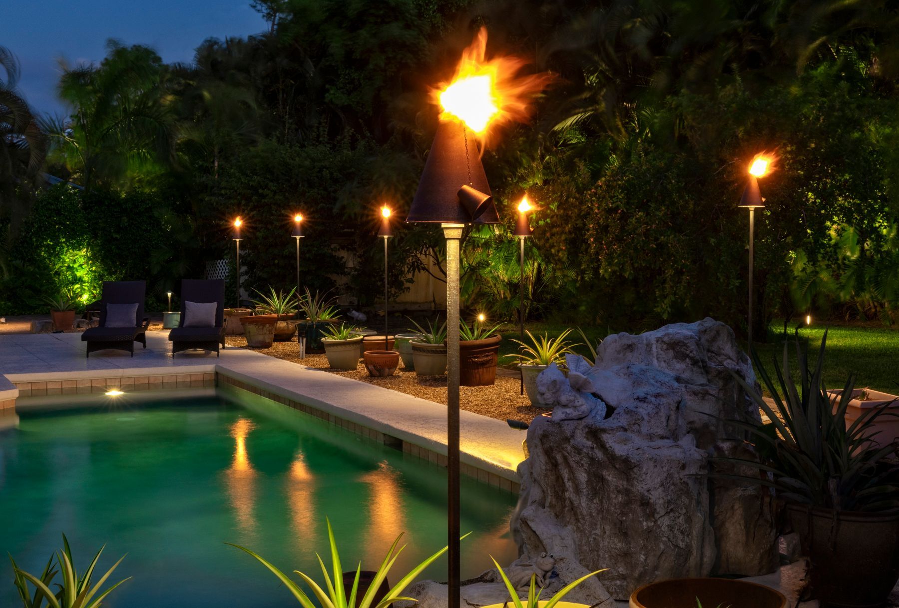 Poolside scene at night, lit by tiki torches and low-level lights. Green pool, trees, and lounge chairs.