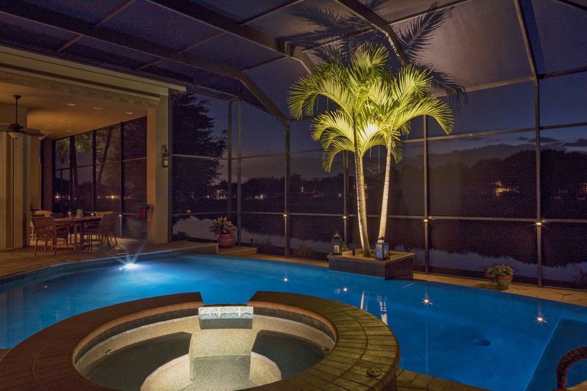 A lit pool and jacuzzi at night, with palm trees and an outdoor dining area visible.