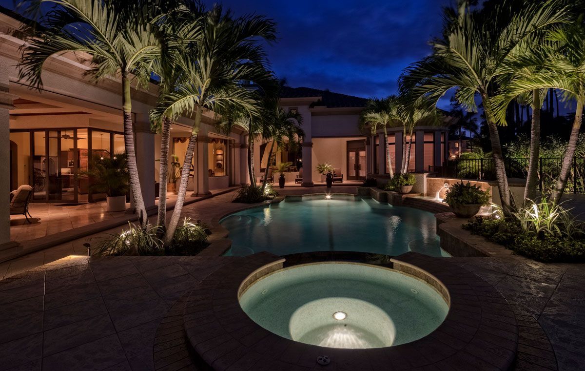 Night view of a luxury backyard with a pool, spa, and illuminated palm trees. A large house is visible in the background.