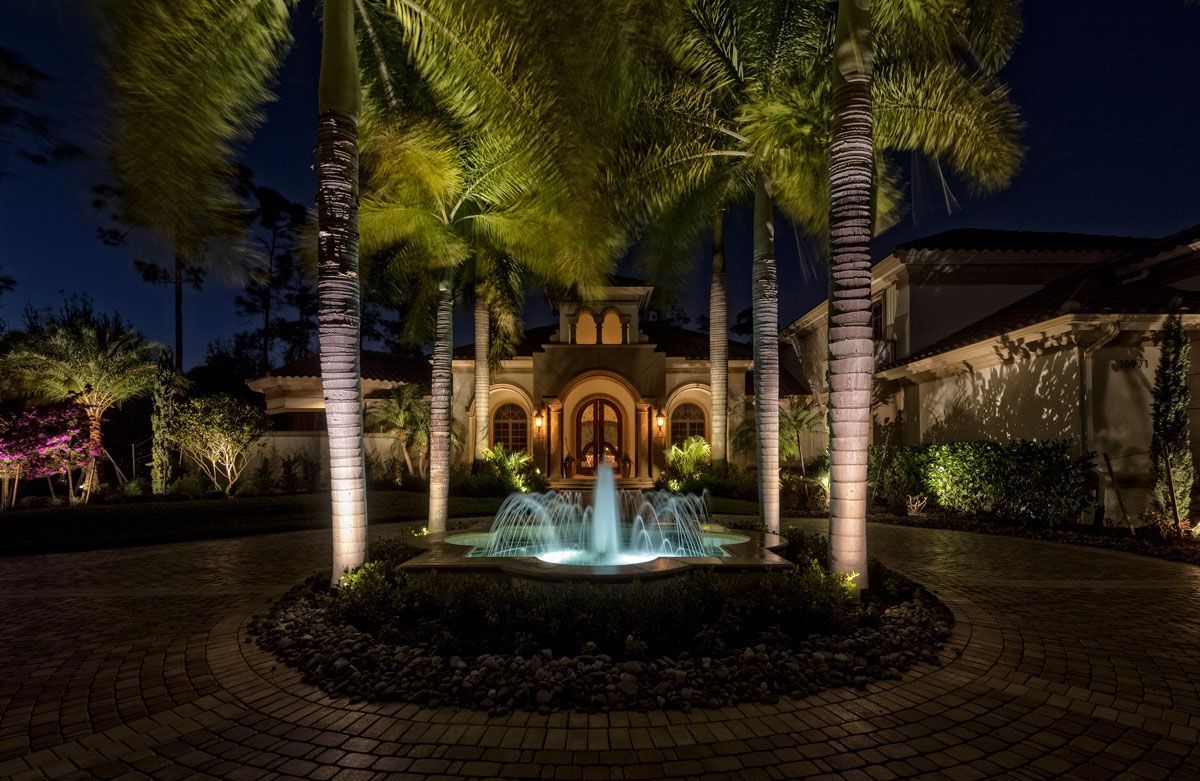 Night view of a luxurious home with a lit fountain, palm trees, and outdoor lighting highlighting the entrance.