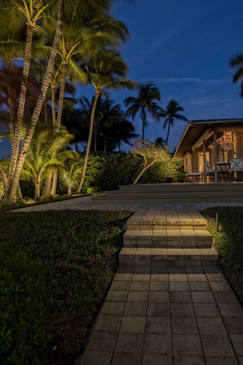 Stone pathway with illuminated steps leading to a building, palm trees in the background at dusk.