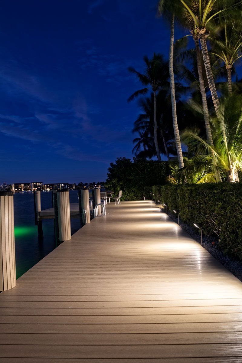 Wooden dock illuminated at night, leading toward the water, palm trees on the right, dark blue sky.