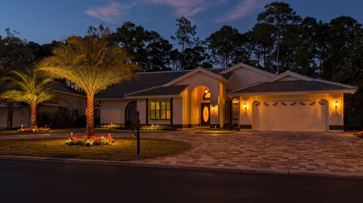 House exterior at dusk, warmly lit with landscaping lights.  Palm trees and front door illuminated.