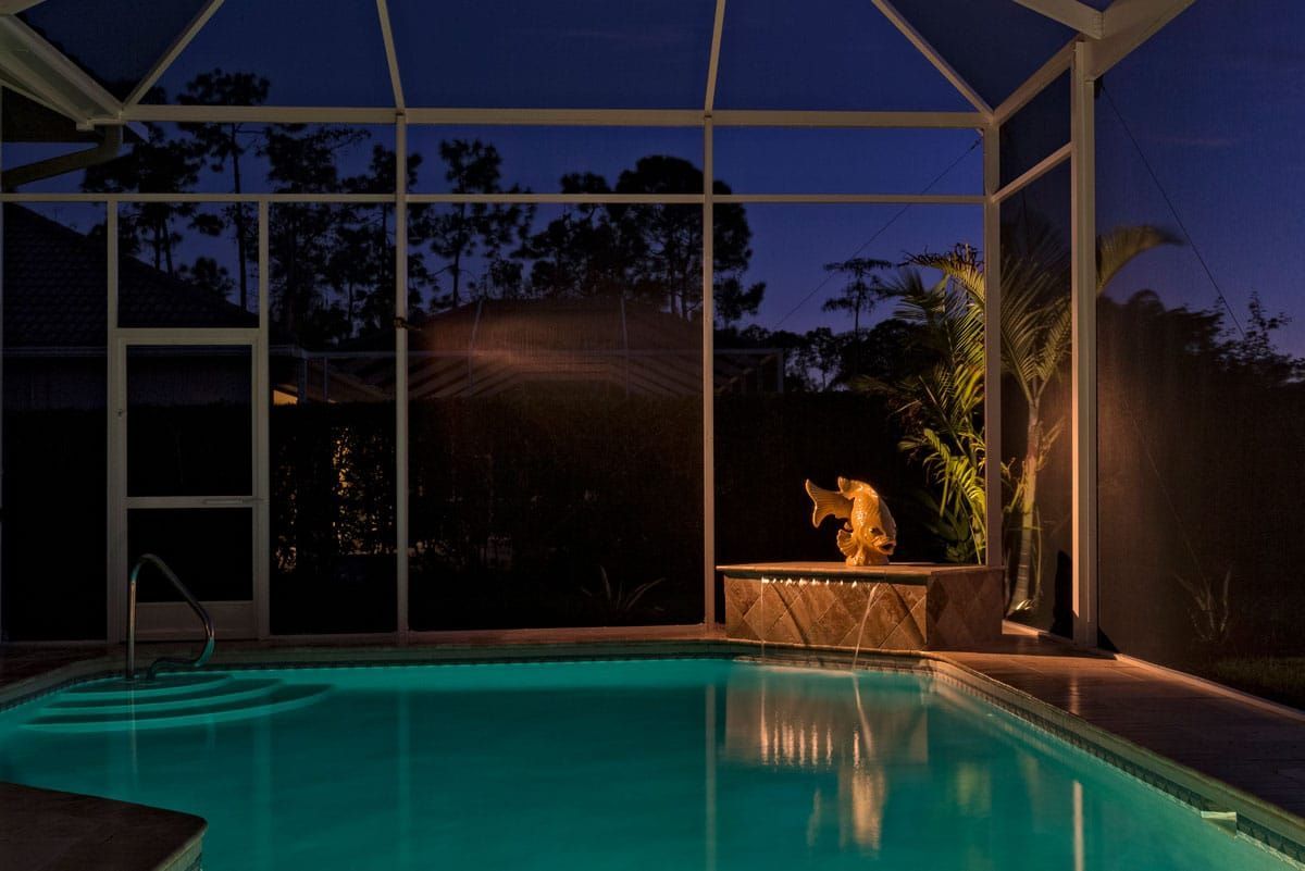 Pool at dusk with illuminated water, framed by a screen enclosure, trees in the background.