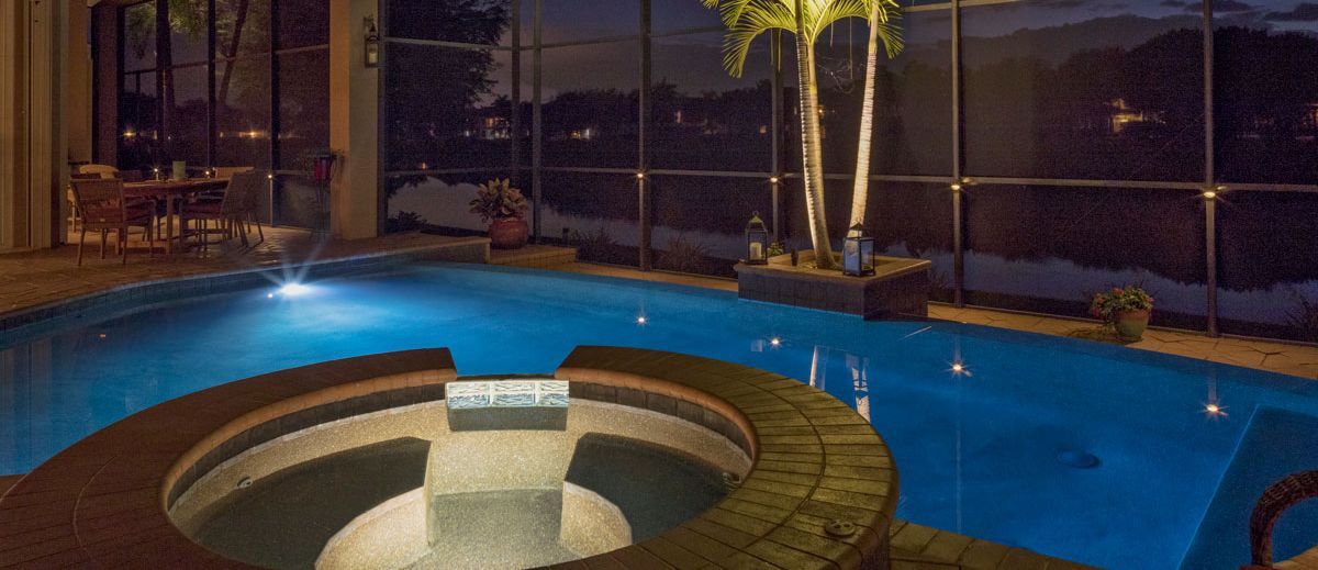 Indoor pool with hot tub, palm trees, and nighttime view through glass panels.