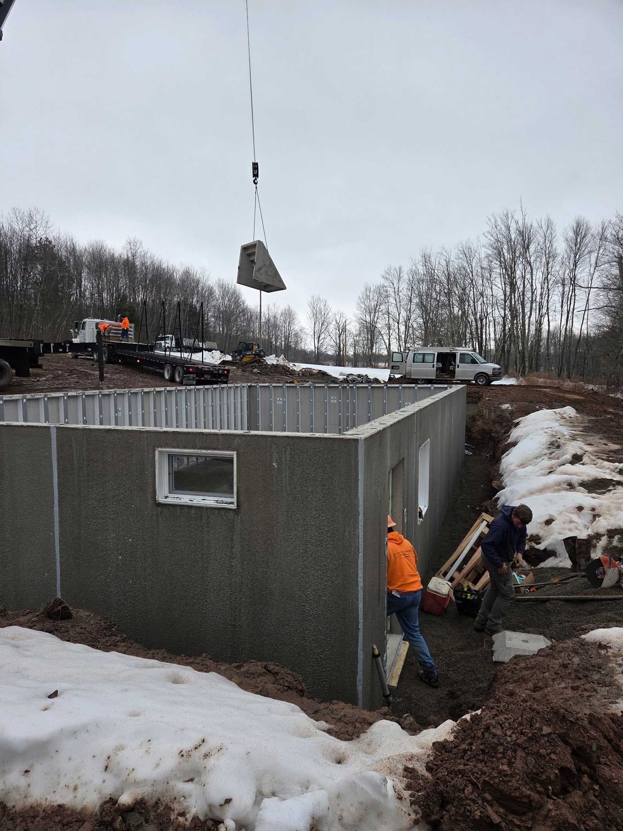 A crane lowers a concrete panel onto a house foundation while construction workers assist in a snowy, wooded area.