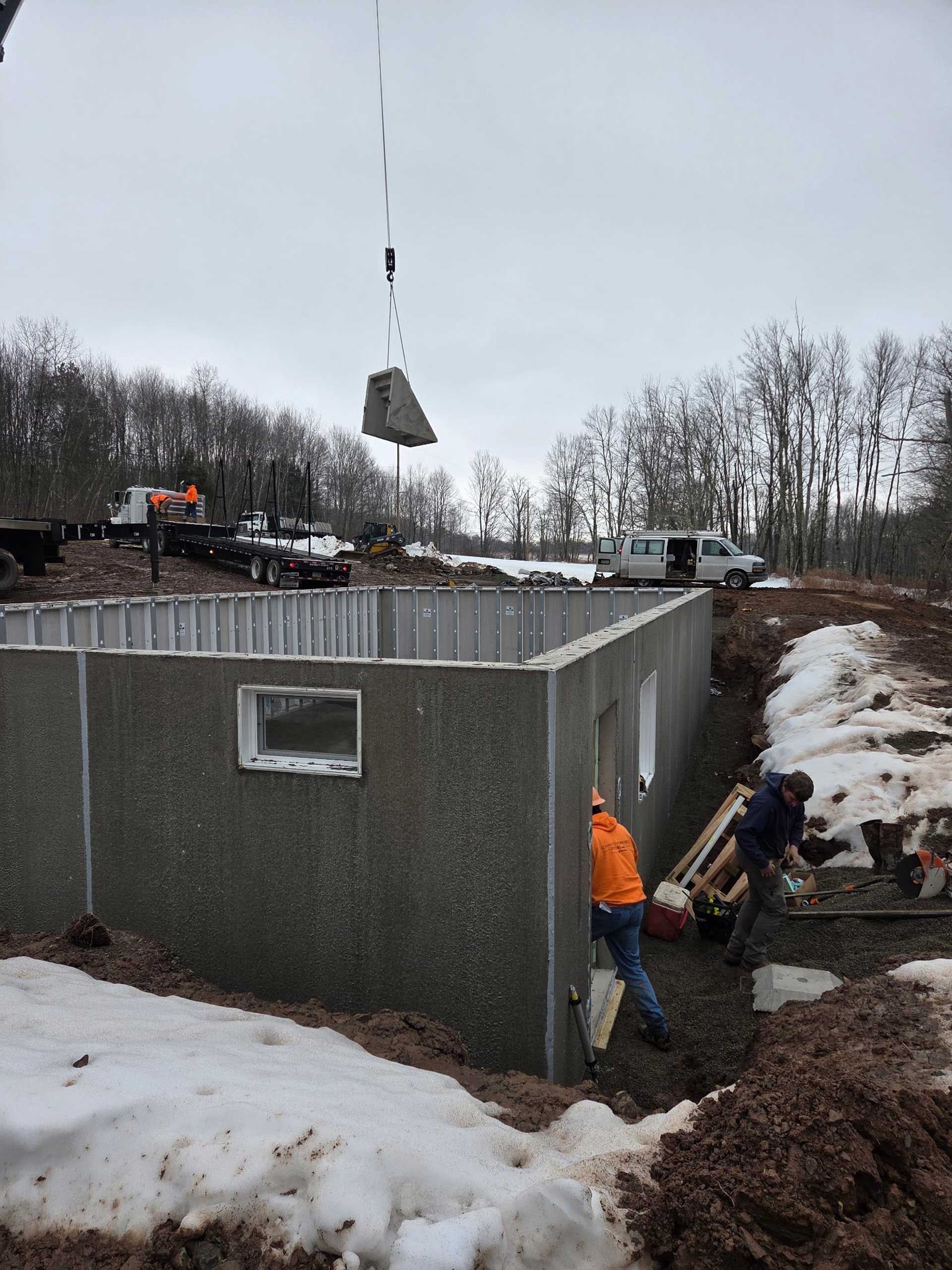 A crane lowers a concrete panel onto a house foundation while construction workers assist in a snowy, wooded area.