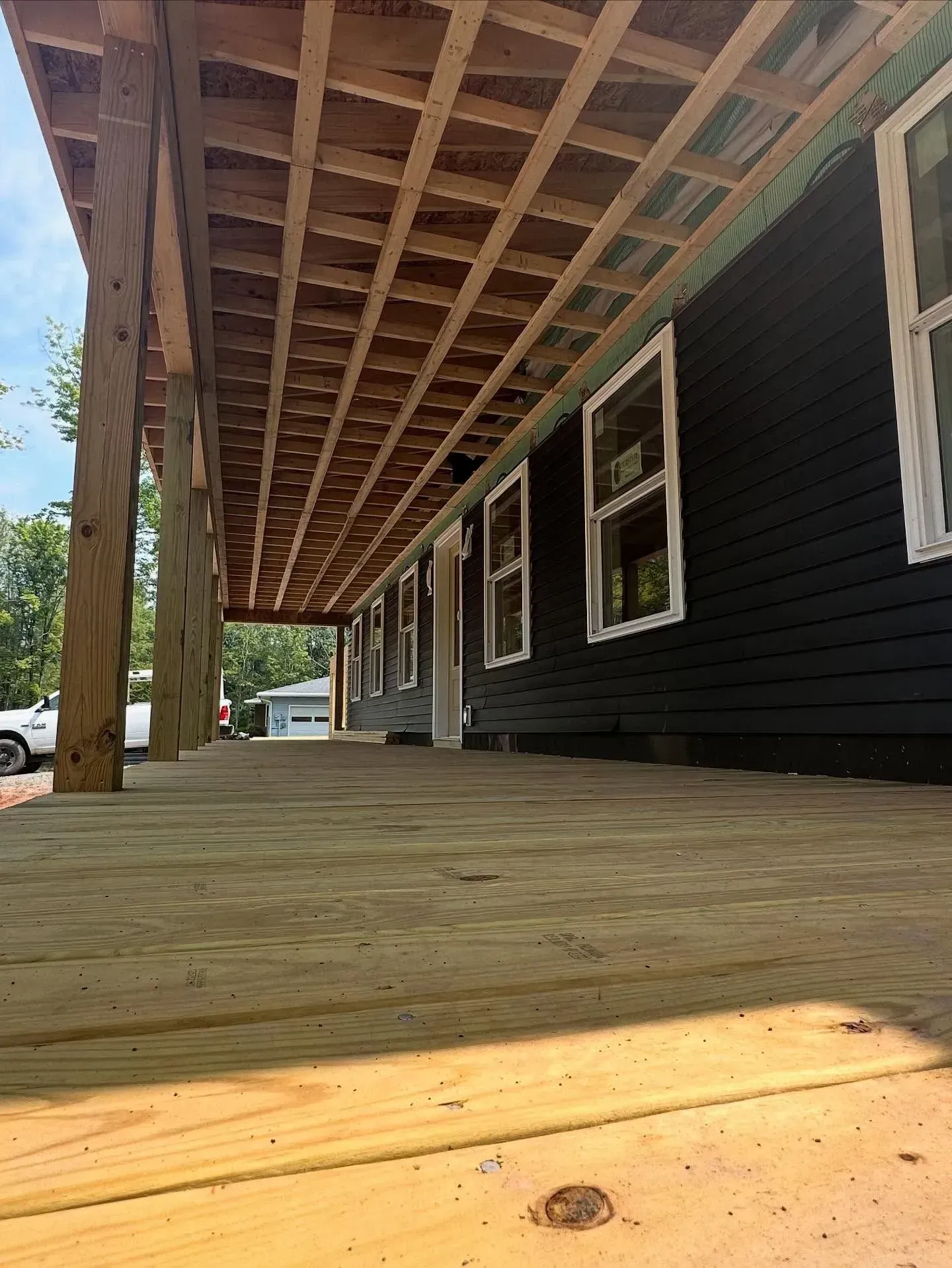A low-angle view of a wooden porch under construction, featuring a newly laid deck, exposed rafters, and dark siding.