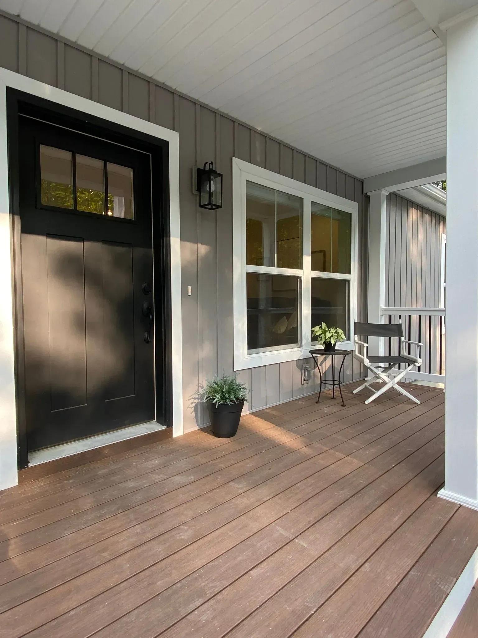 A front porch with gray vertical siding, a black door with window panes, a potted plant, and a folding chair.