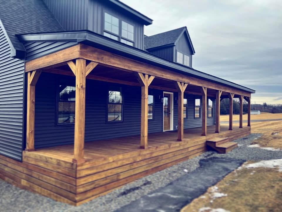 A dark-sided farmhouse features a wrap-around wooden porch with timber posts and a metal roof against an overcast sky.