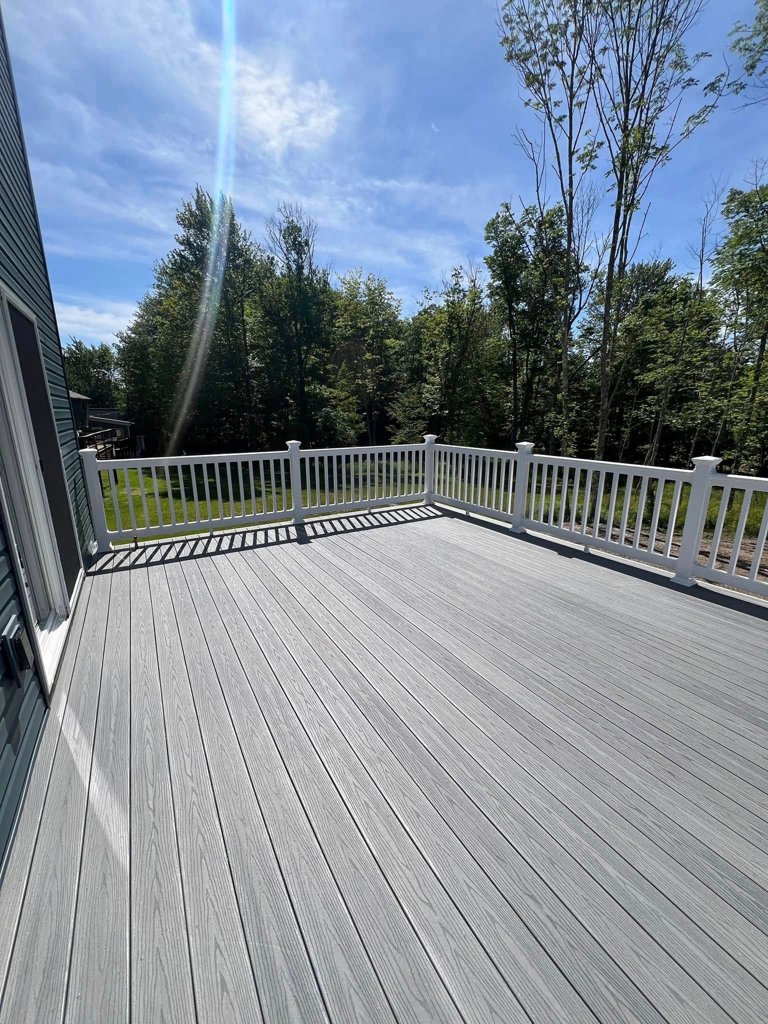A grey composite deck with a white railing overlooks a lush, sunlit forest under a bright blue sky.