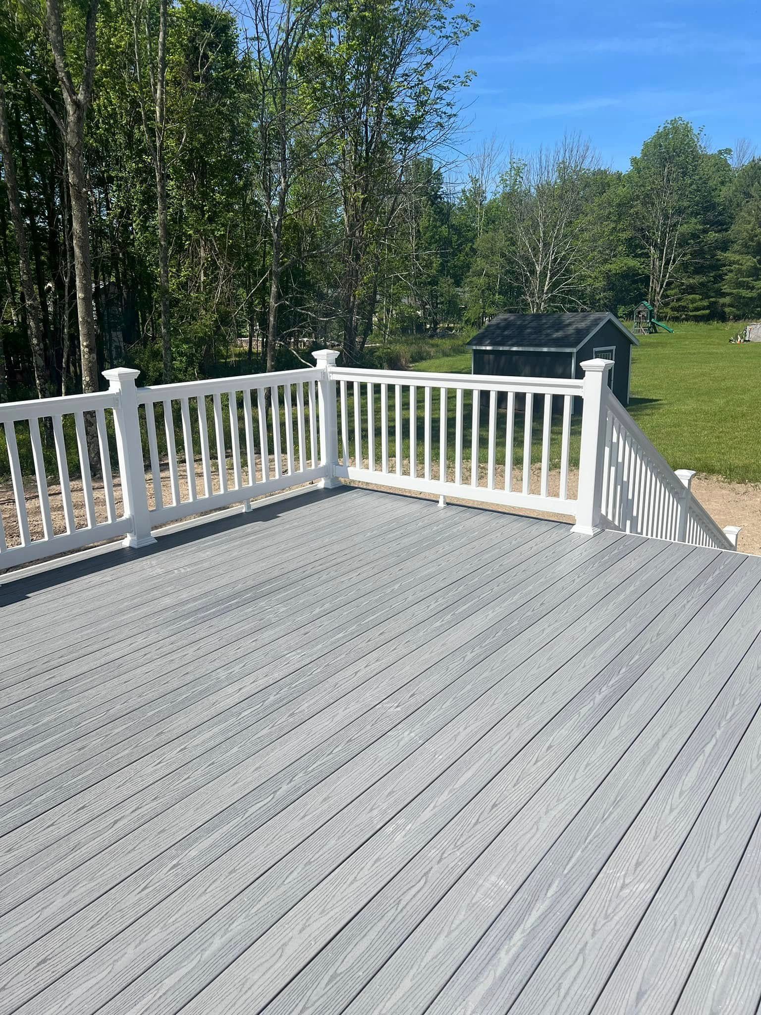 A grey composite deck with white railings, set against a backdrop of green trees and a small storage shed under a blue sky.