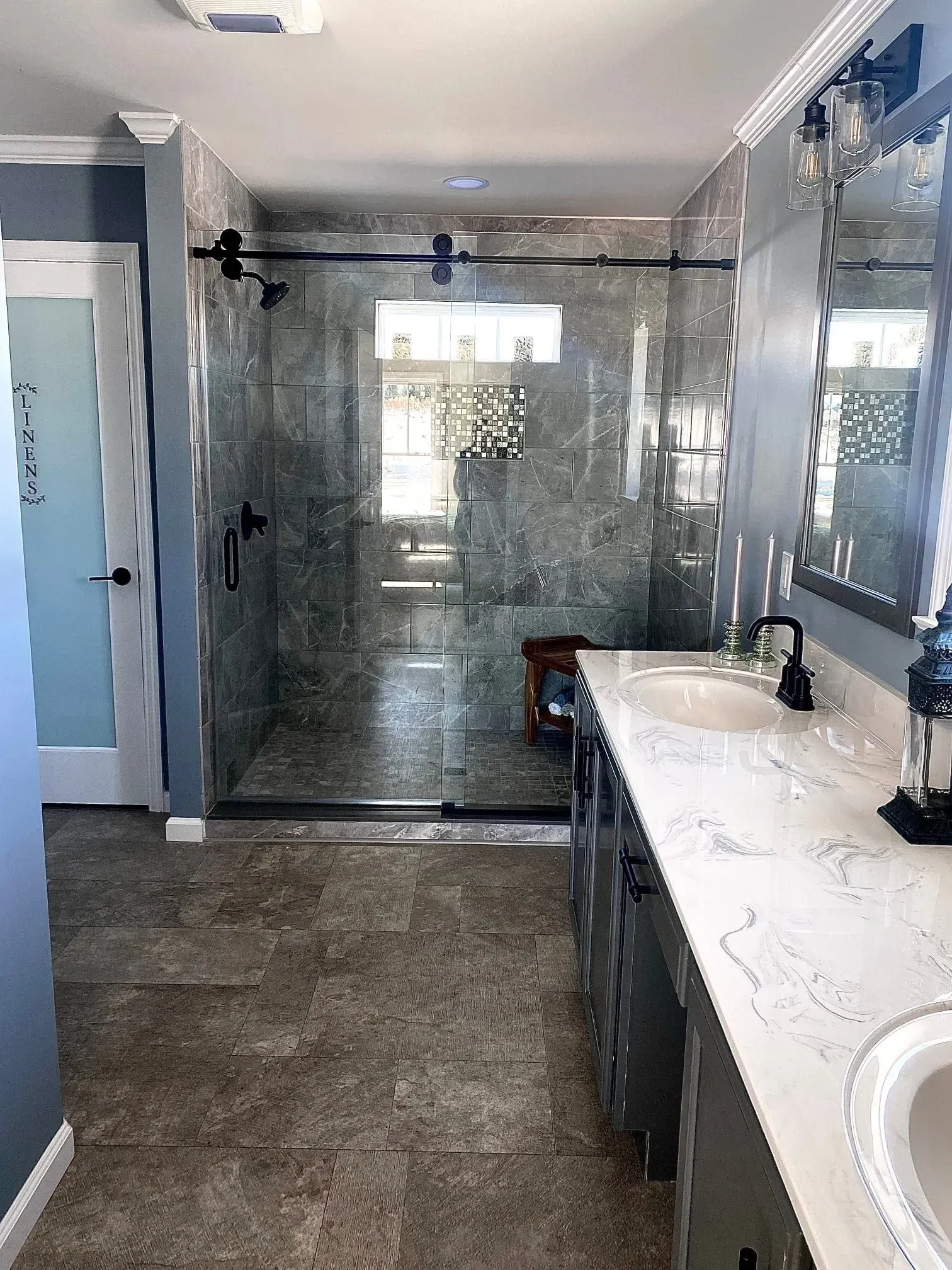 A bathroom featuring a walk-in glass shower, a marble-topped vanity with gray cabinets, and dark stone floor tiles.