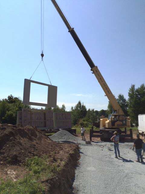 Crane lifting a concrete wall panel during building construction on a gravel driveway.