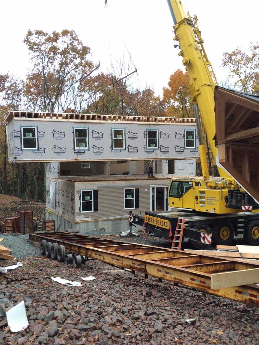 A crane lifting a modular home's second story into place; construction site with trees in the background.