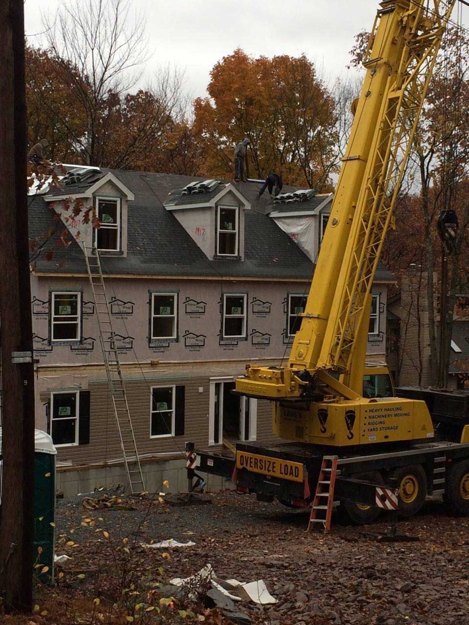 House under construction, workers on roof, yellow crane alongside. Brown siding, cloudy sky, fall trees.