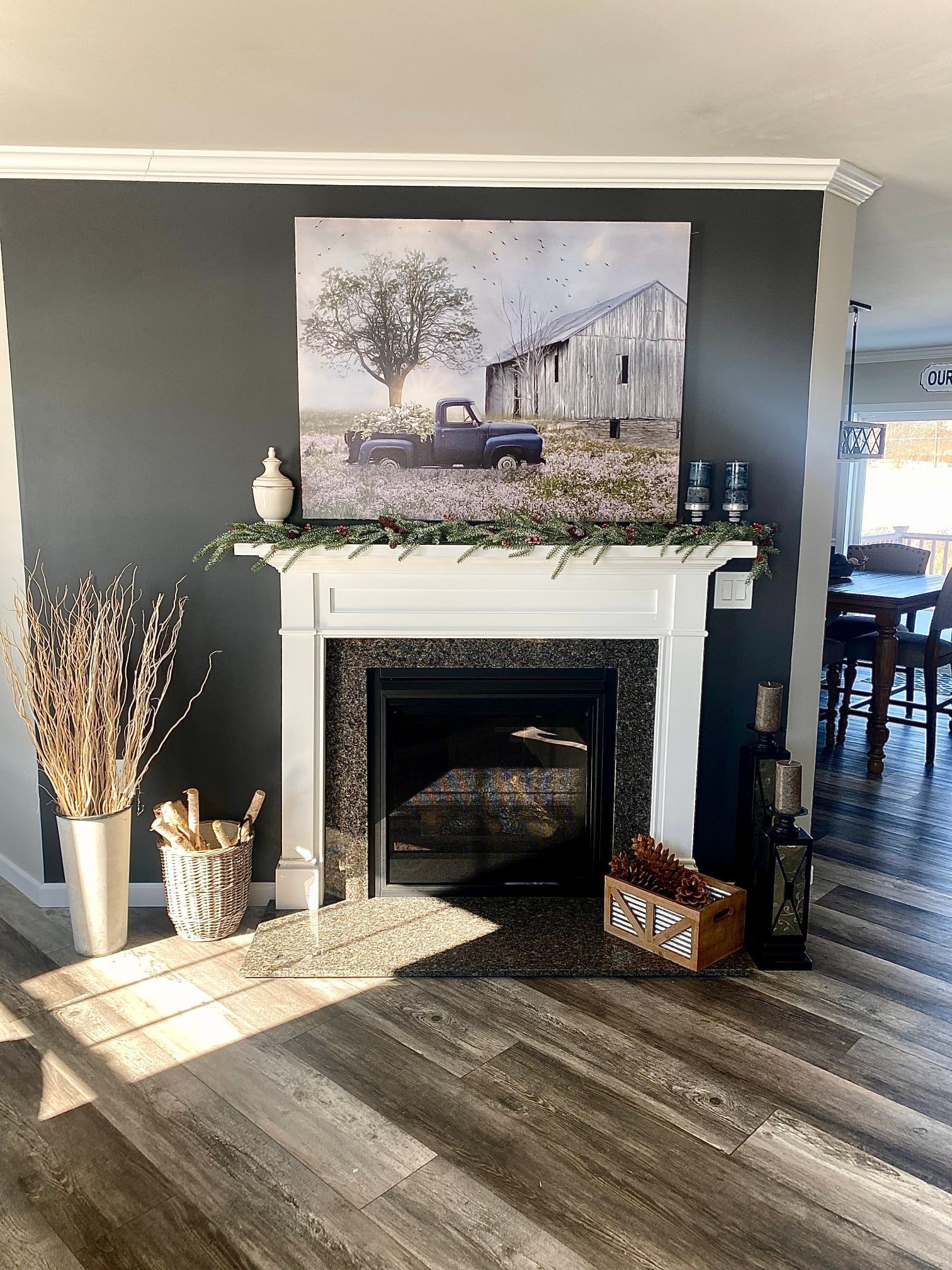 Fireplace with artwork of a barn and truck, decorated with greenery and dried branches; wooden floor.