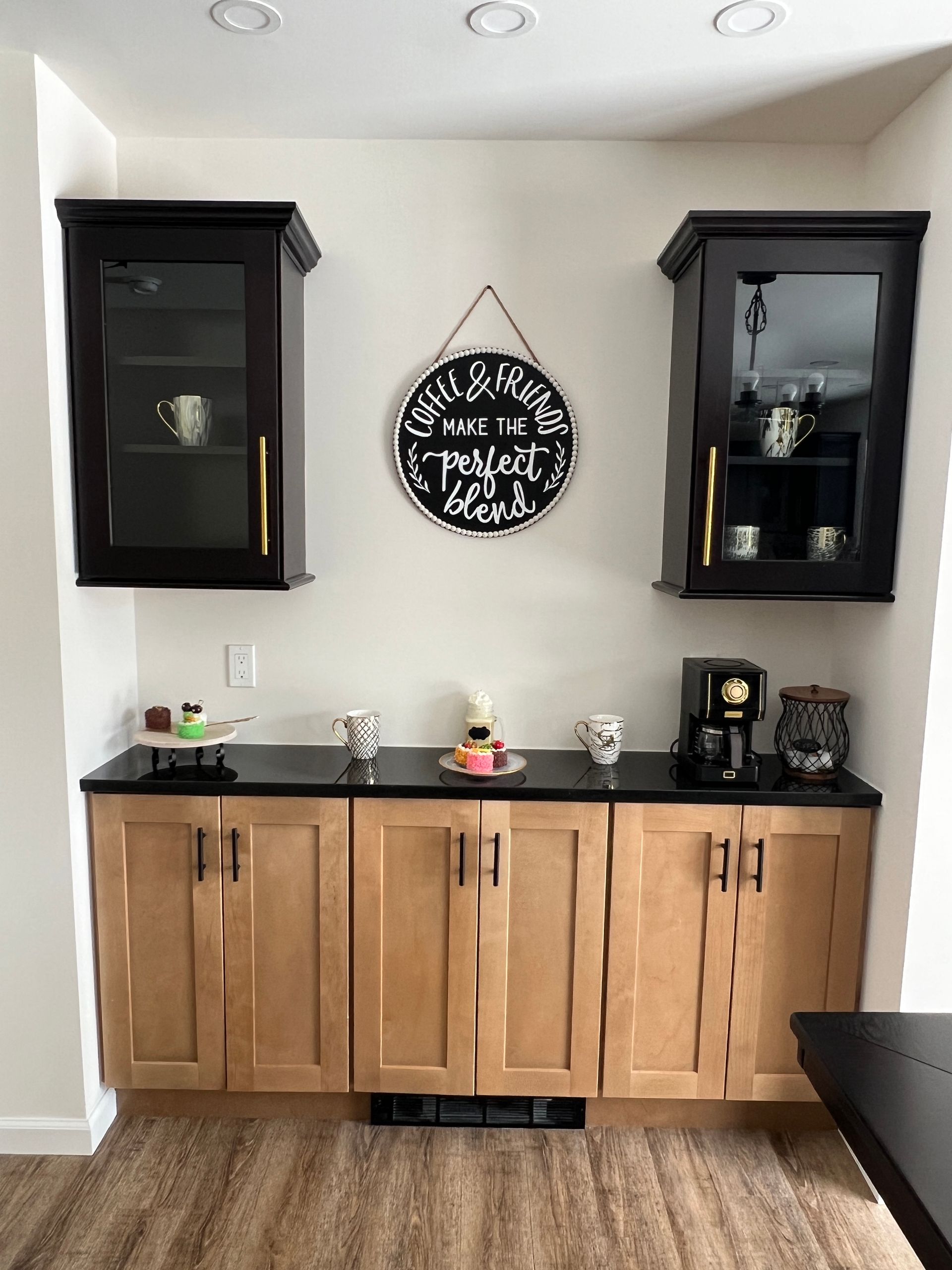 Coffee station with black countertop, light cabinets, black cabinets, and a sign.