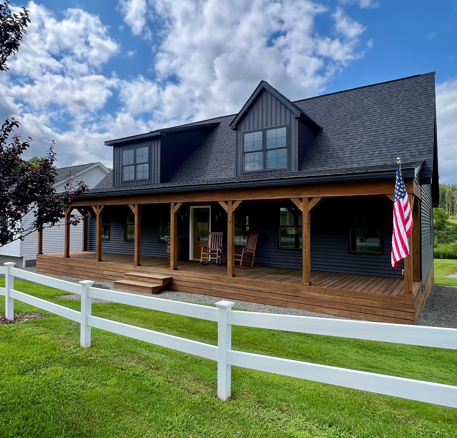 Dark gray farmhouse with wooden porch, rocking chairs, and American flag on a sunny day.