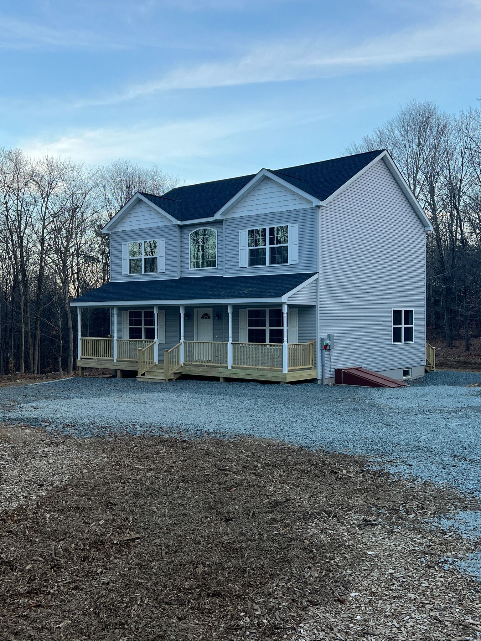 Two-story house with light blue siding, black roof, and wrap-around porch in a gravel-covered yard.