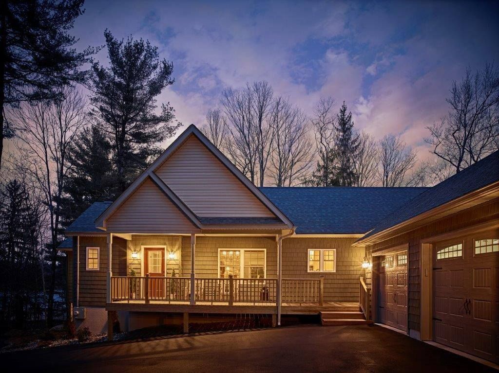 Cottage-style home at dusk with lights on, front porch, attached garage, set in wooded area.