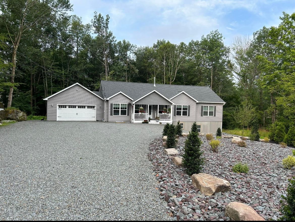 A gray, ranch-style house with a gravel driveway, surrounded by trees.