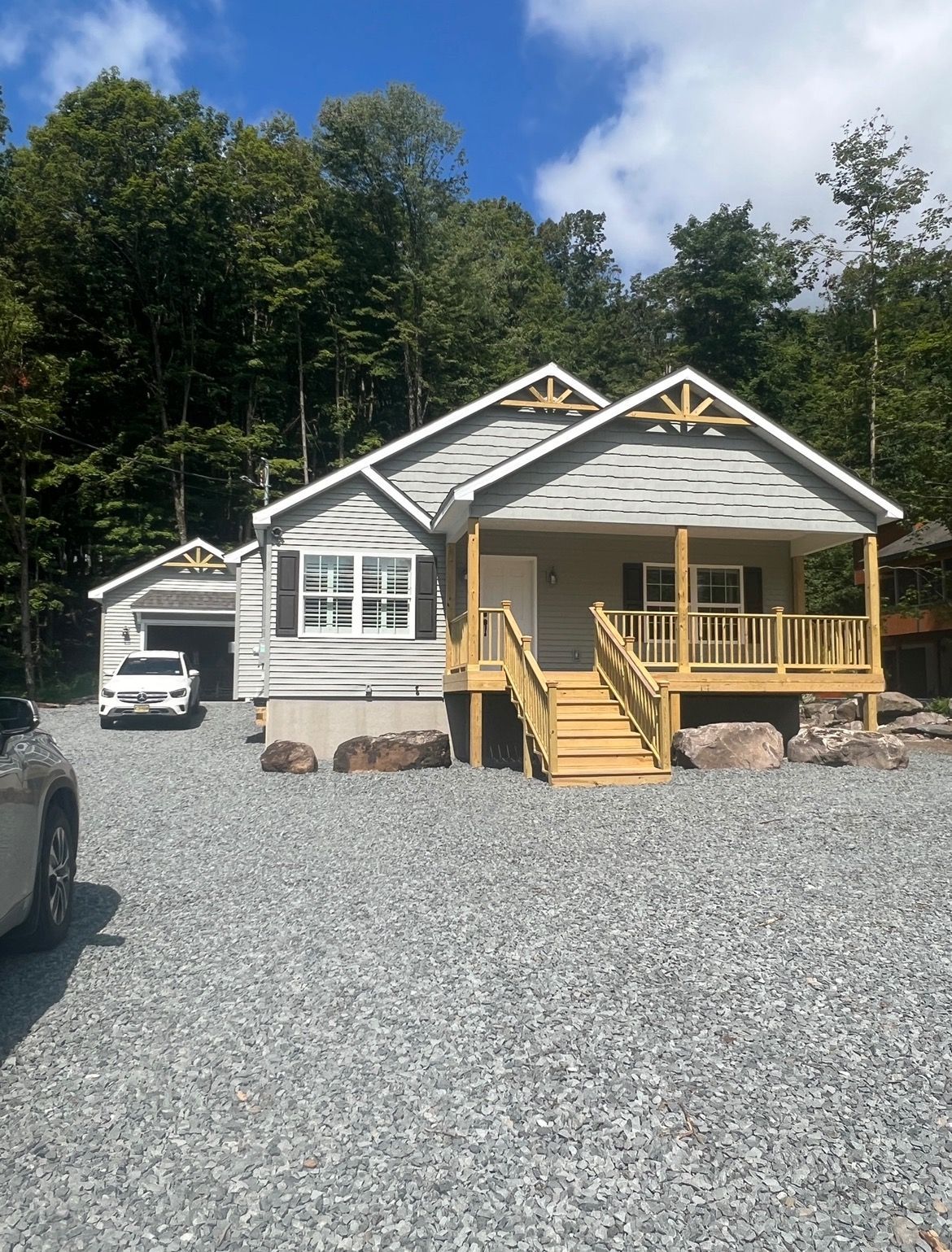 A light-colored house with a porch and garage on a gravel driveway, surrounded by trees.
