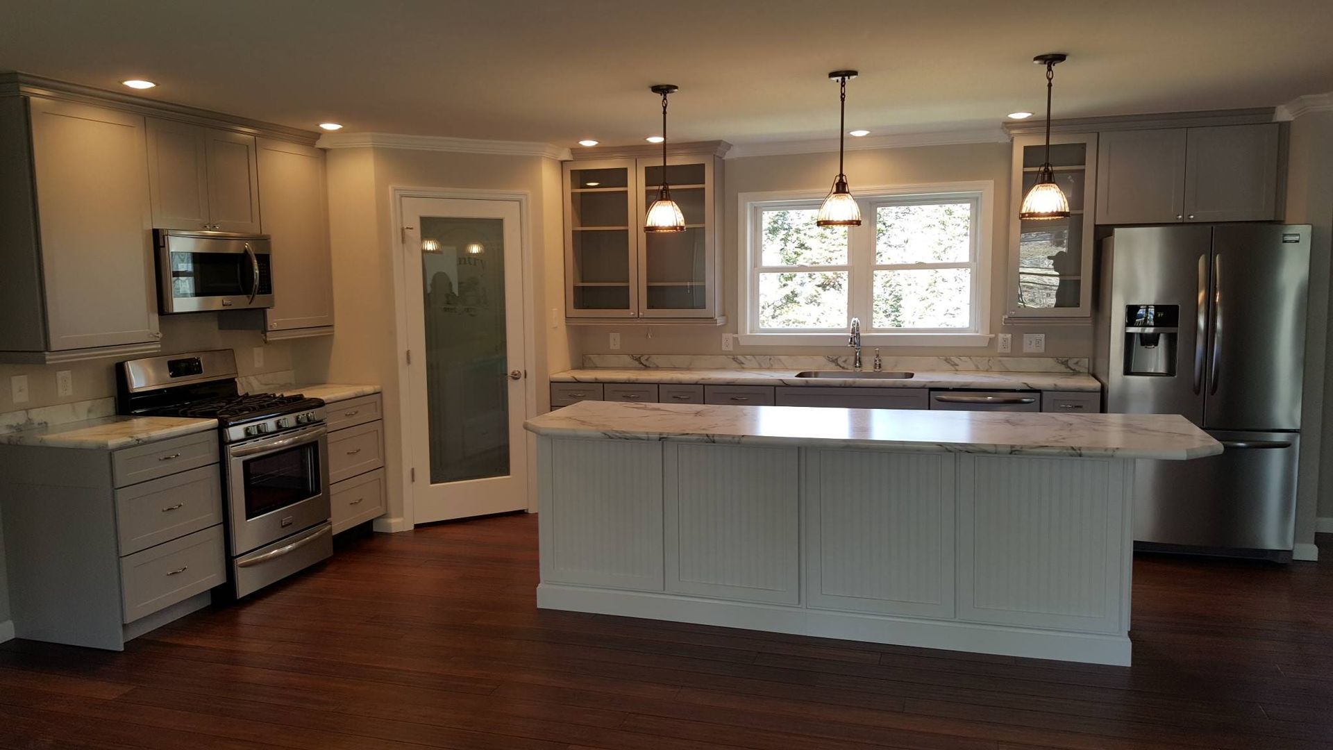 A modern kitchen featuring white cabinets, stainless steel appliances, a marble-topped island, and dark hardwood floors.