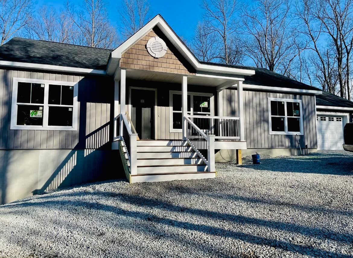 A single-story gray house with a front porch, wood-shingle gable, and gravel driveway set against bare trees under blue sky.