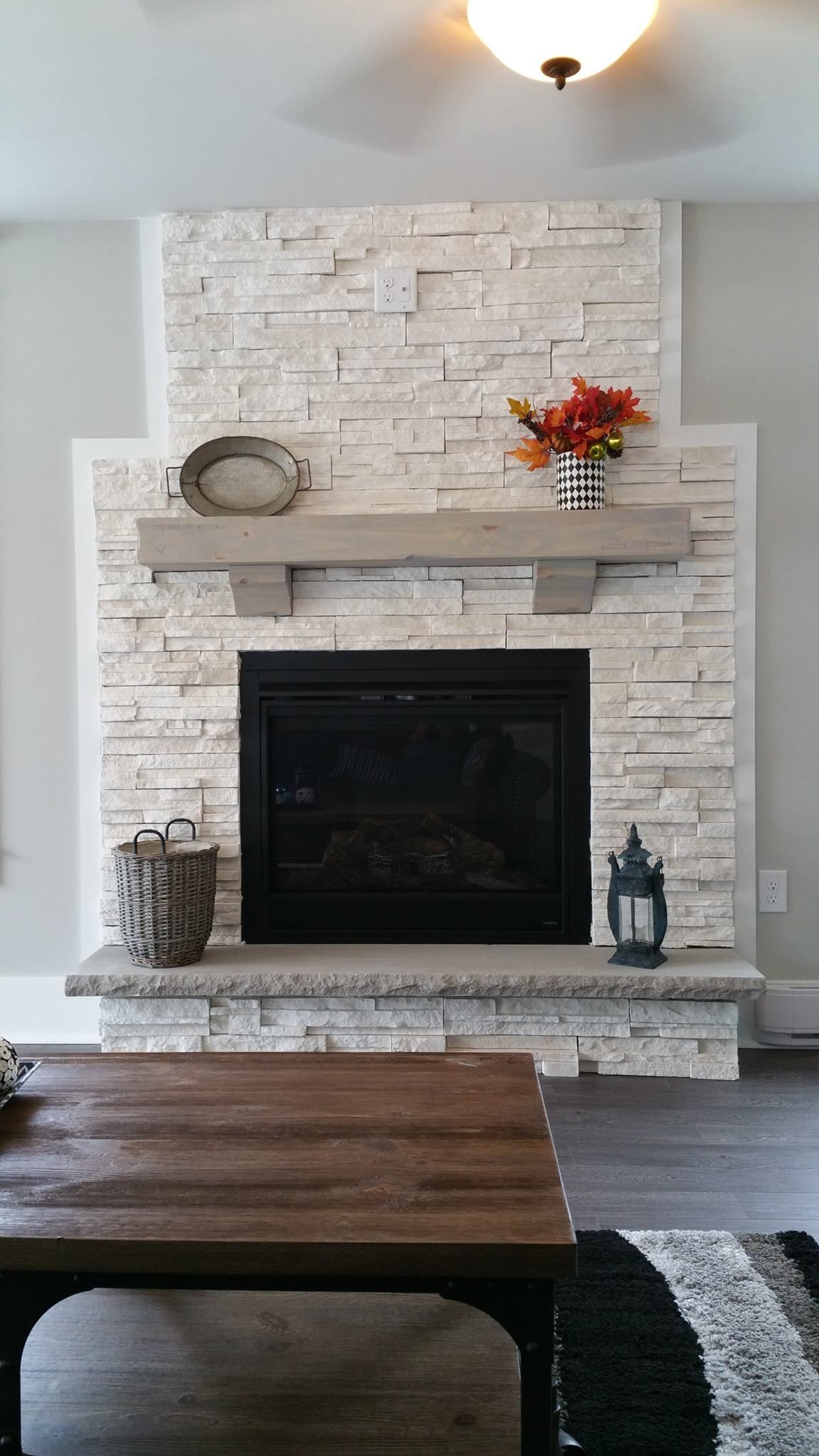 A white stone fireplace with a thick wooden mantel, a black hearth, and decor, viewed from a rustic living room.