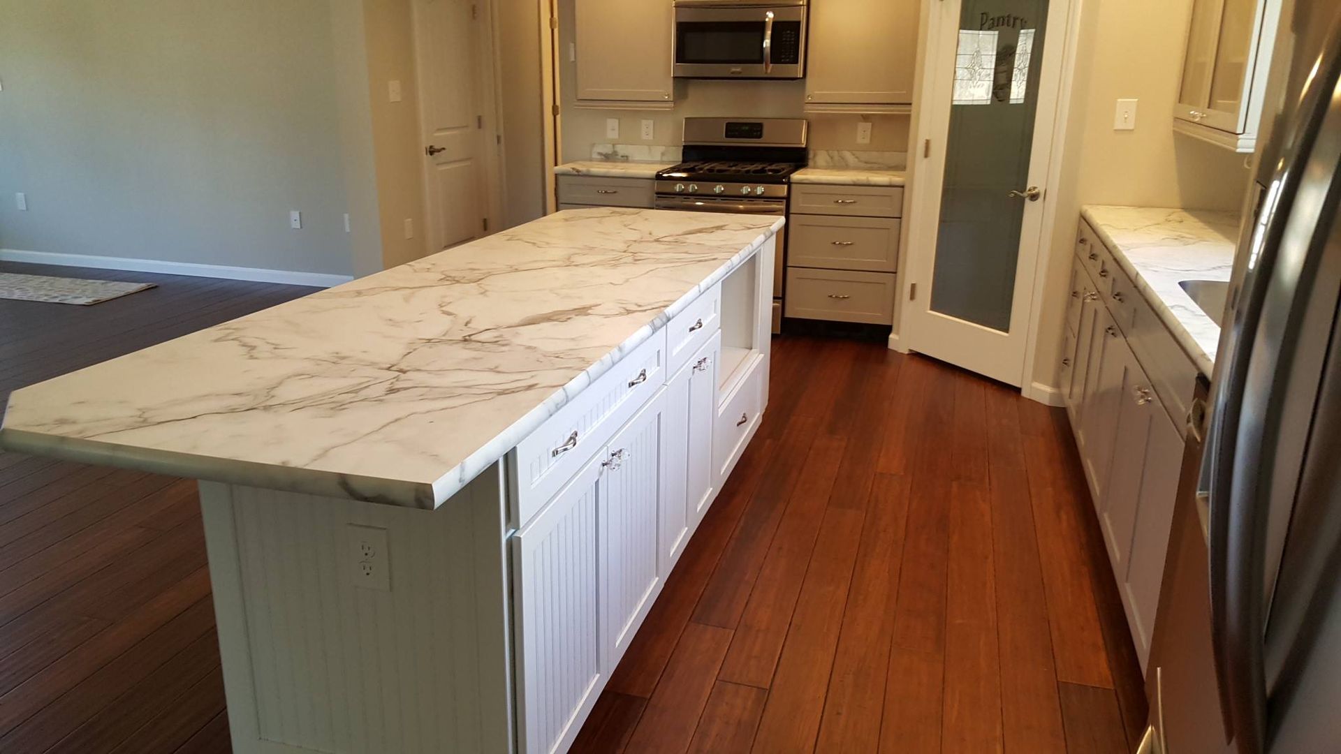 A kitchen island with a white marble countertop sits in front of light cabinets, a stove, and a glass-paneled pantry door.
