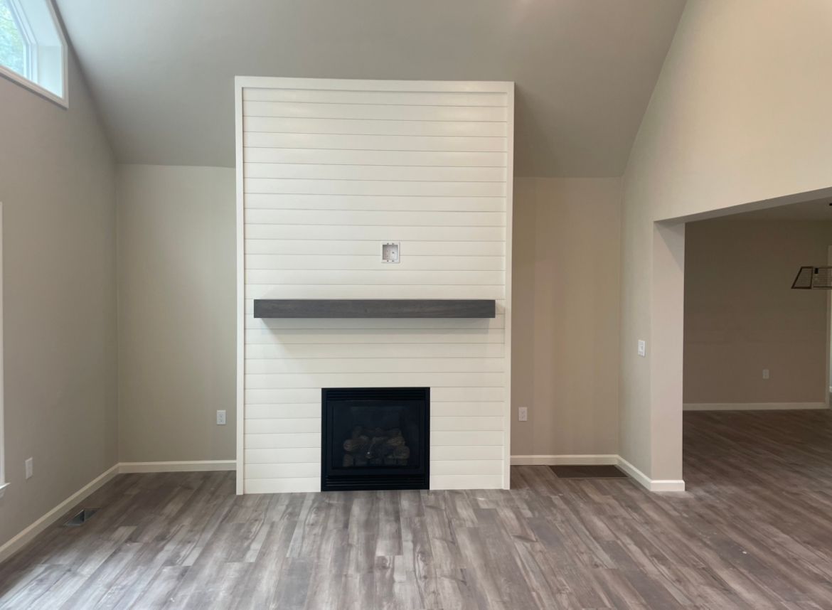 A white shiplap fireplace with a dark grey floating mantel sits in a room with grey wood flooring and neutral walls.