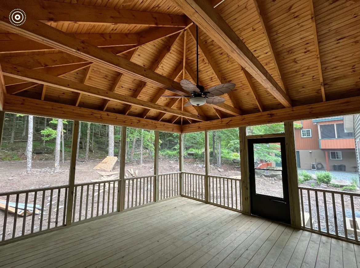 A screened-in wooden porch with vaulted ceilings and a ceiling fan, overlooking a wooded yard and a nearby house.