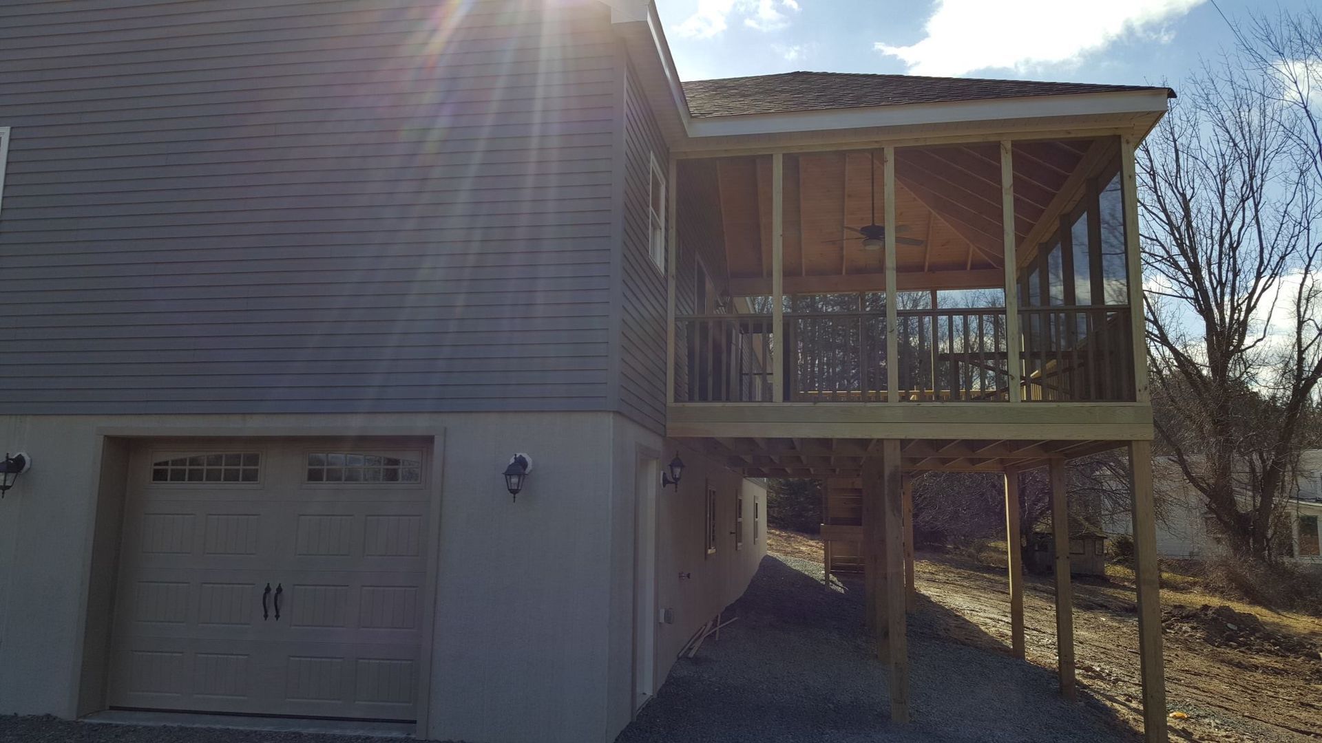 A house exterior featuring a beige basement garage and an elevated screened-in wooden porch on a bright, sunny day.