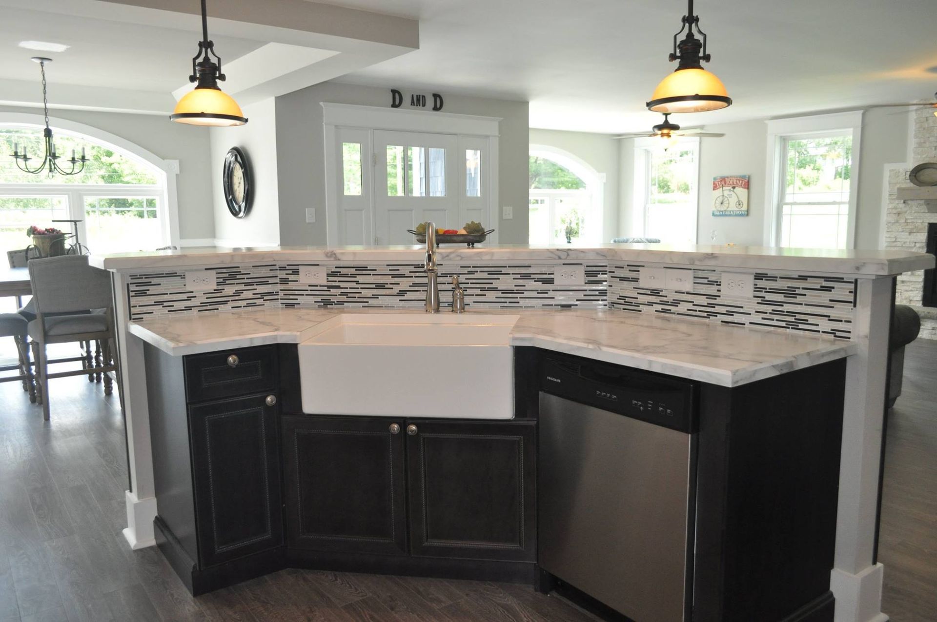 A kitchen island with a farmhouse sink, dark cabinets, marble countertops, and a decorative tile backsplash.