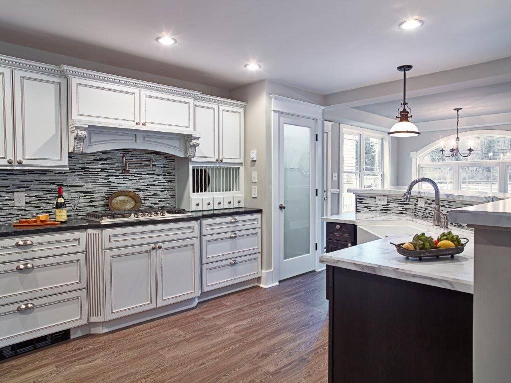 A kitchen with white cabinets, dark island, marble countertops, stone backsplash, and wood-style flooring.