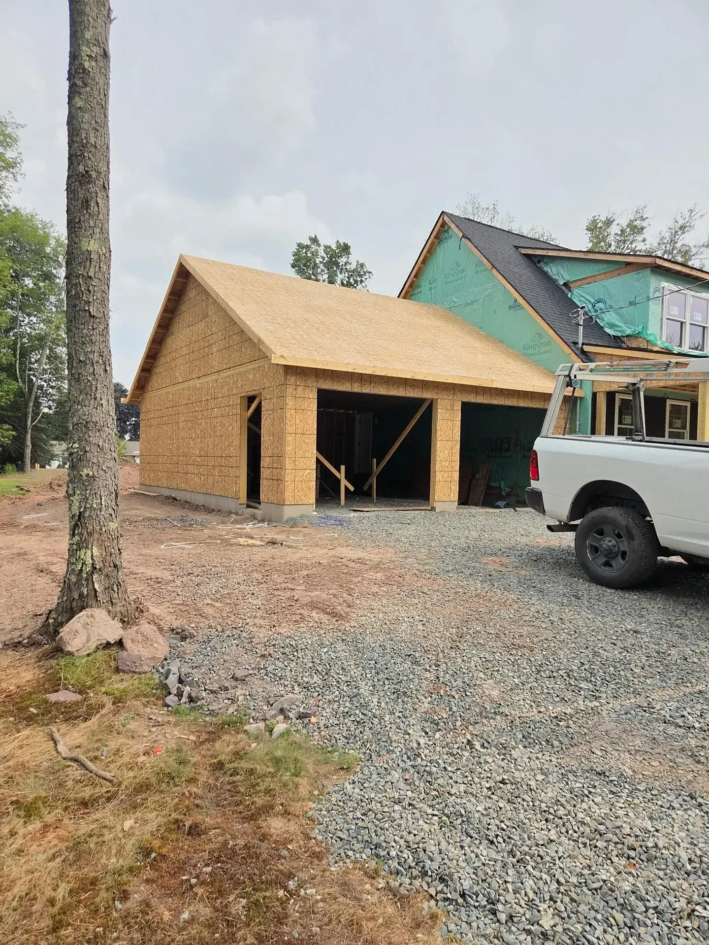A newly constructed wooden garage frame stands next to a house under construction, with a white truck parked nearby.