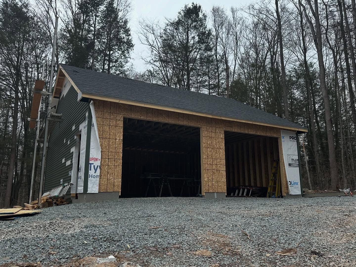 A two-car garage under construction with plywood siding and a grey shingled roof, surrounded by a forest.