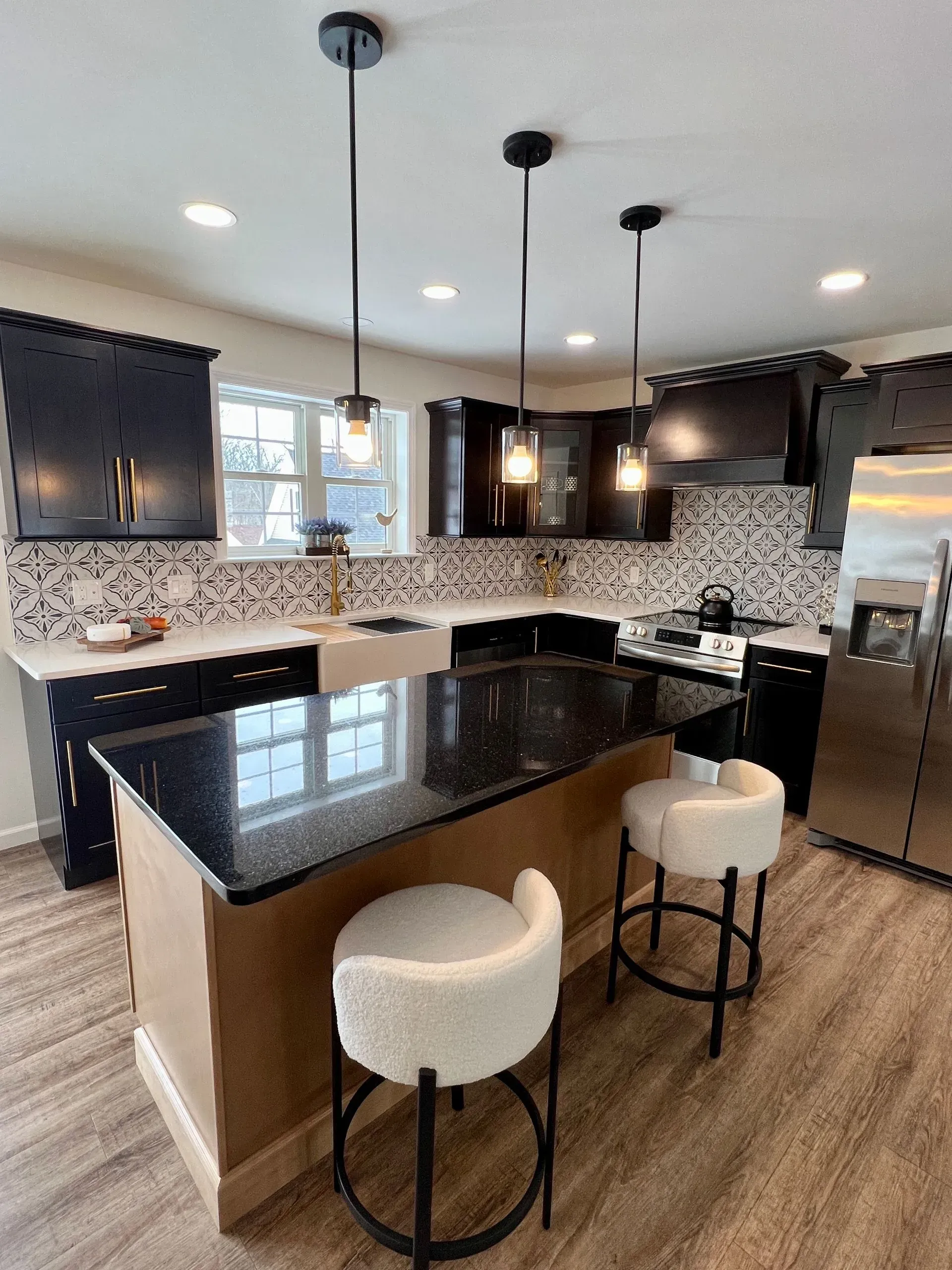 A modern kitchen with black cabinets, a granite island, white patterned backsplash, and two beige stools under pendant lights.