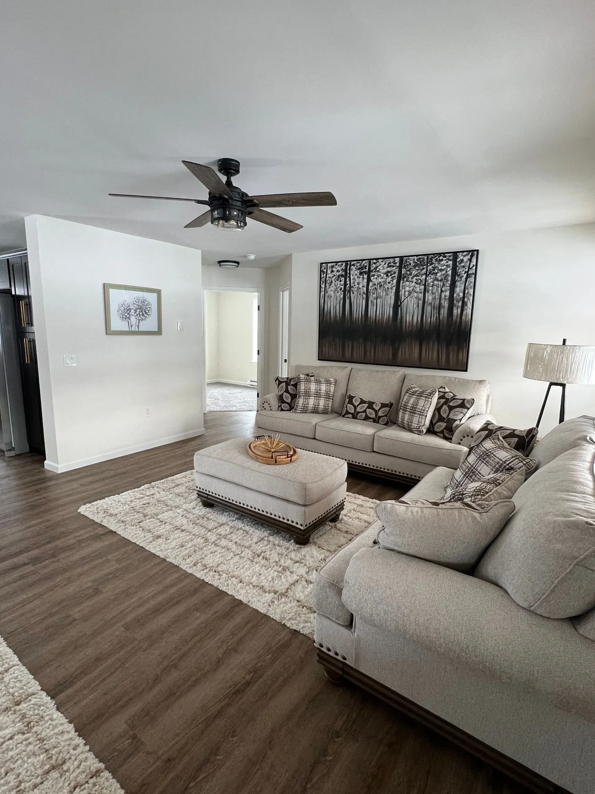 A bright, modern living room featuring a light gray sofa and ottoman, dark flooring, and a large black-and-white tree mural.