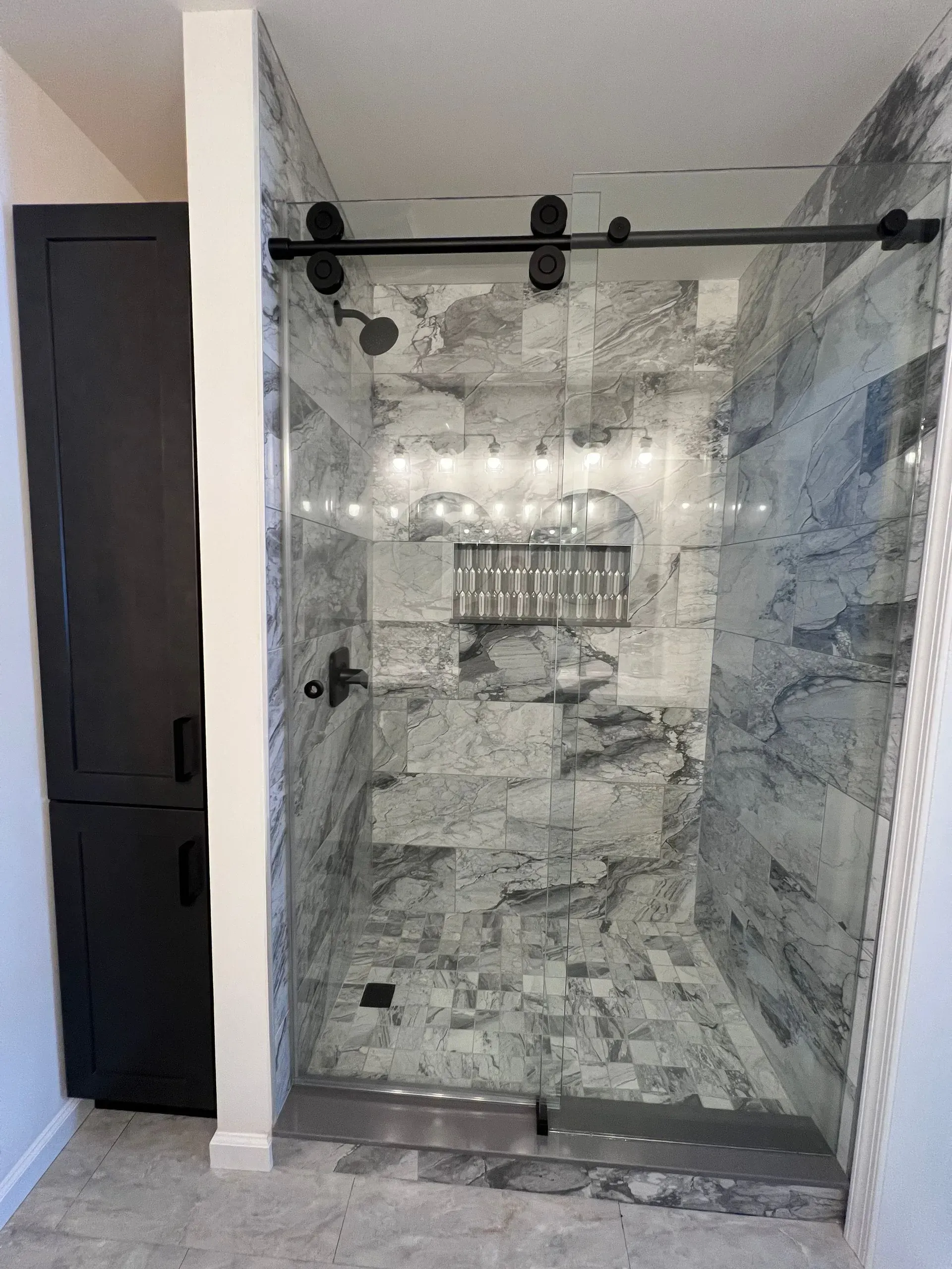 A walk-in shower with grey and white stone-patterned tiles, a black sliding glass door, and a tall, dark wood cabinet.