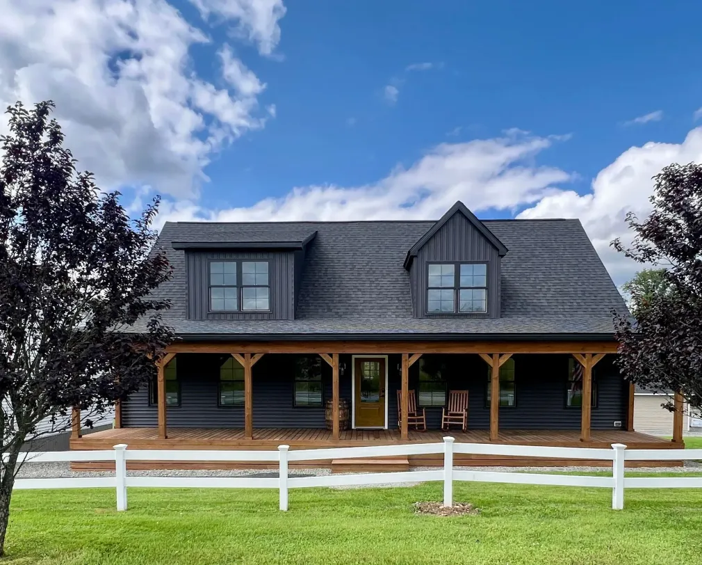 A dark charcoal house with a wood porch and trim, two dormer windows, and a white fence in front under a cloudy blue sky.