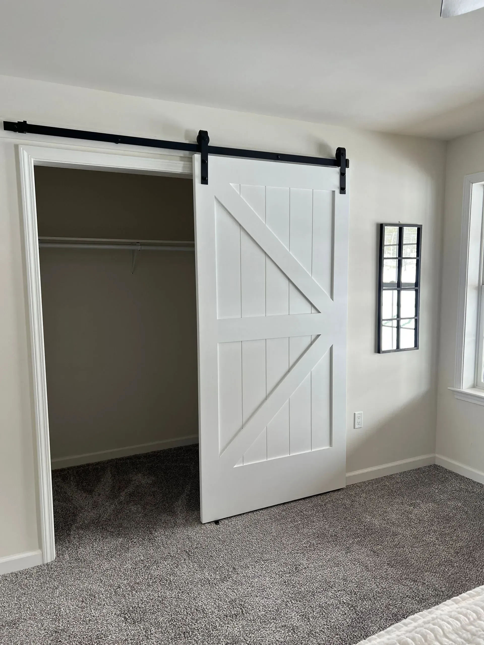 A white sliding barn door on a black rail hangs in front of an open closet in a bedroom with grey carpet.