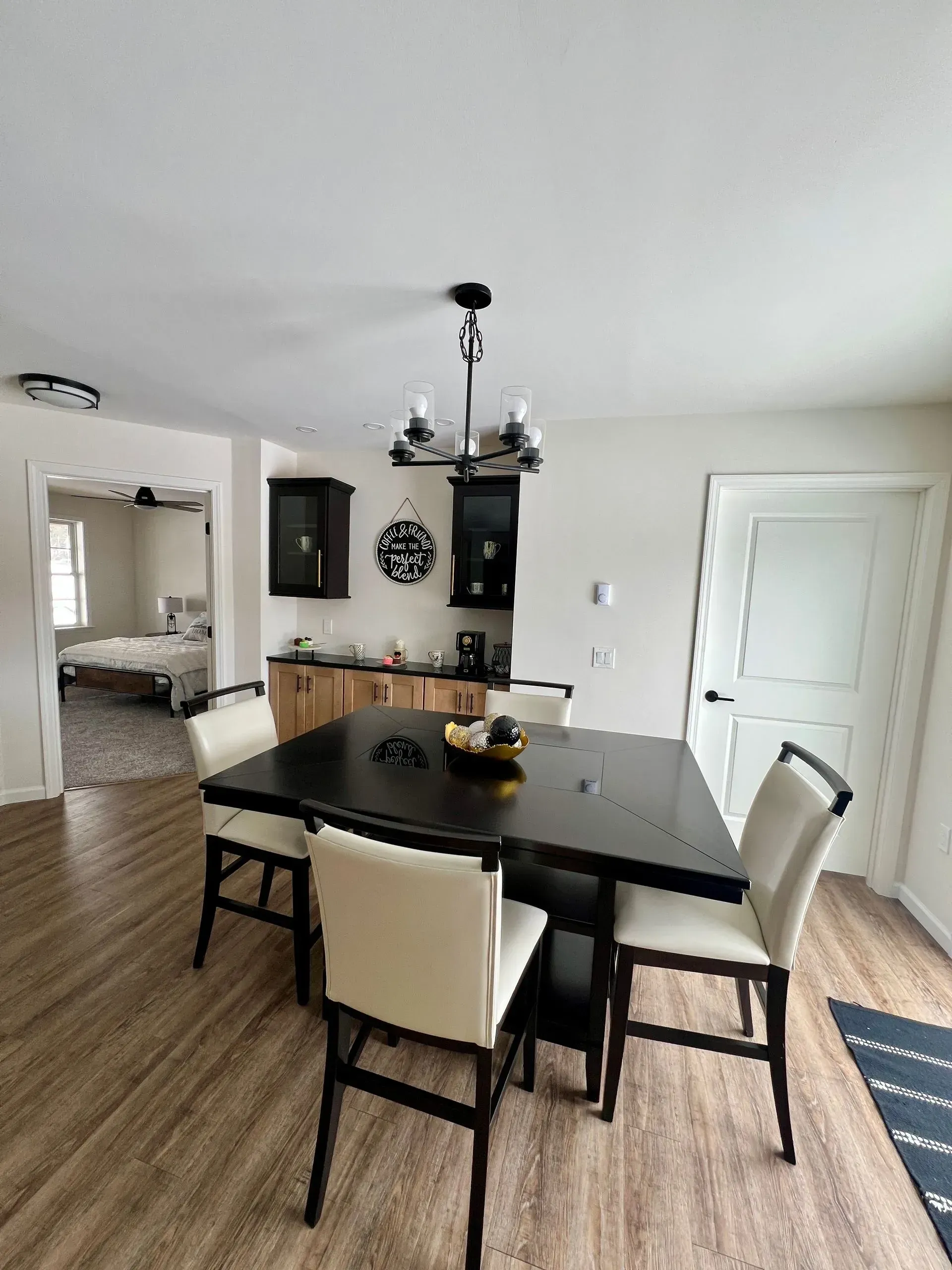 A dining area with a black square table, four off-white chairs, a dark wood sideboard, and a chandelier.