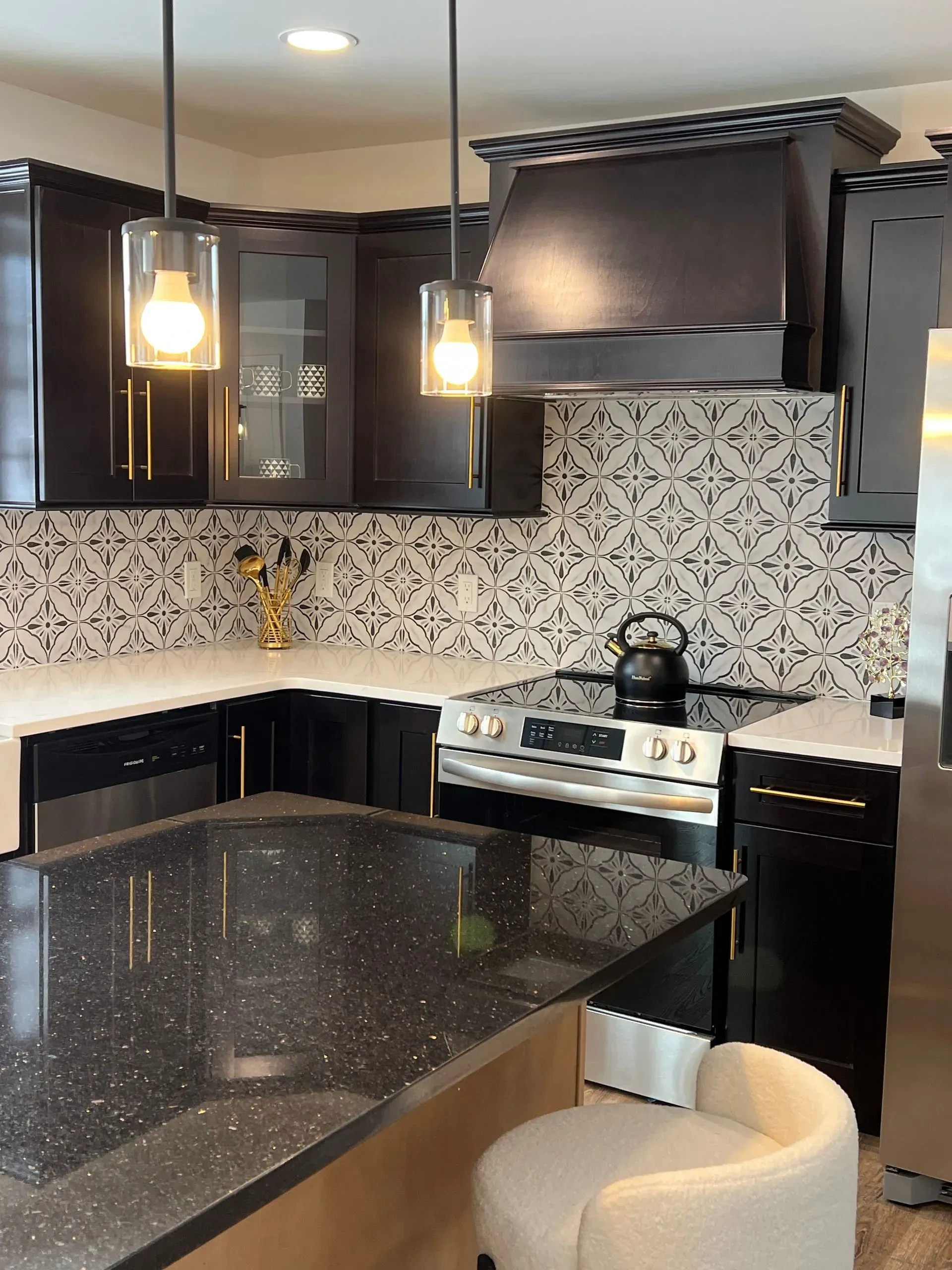 A kitchen features dark cabinetry, patterned white and black backsplash tiles, black stone countertops, and pendant lights.